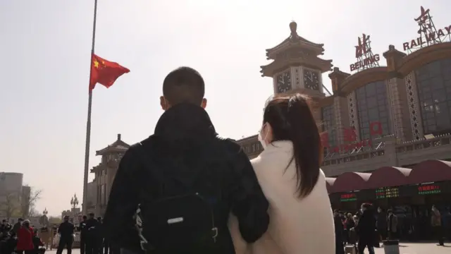 A couple pauses in front of a flag at half-mast in Beijing