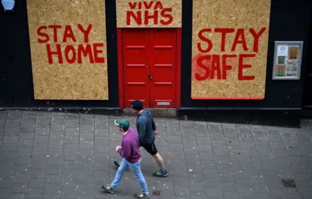 Two pedestrians walk past a 'Stay at home, stay safe' sign