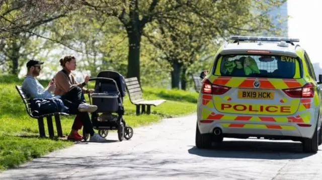 A police car drives past people sitting on a bench