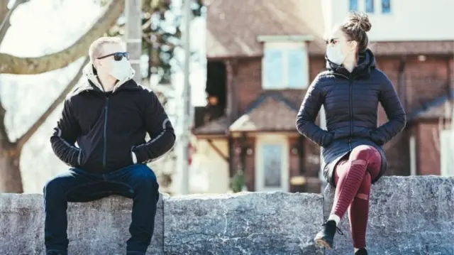 Two people wearing masks sit on a bench