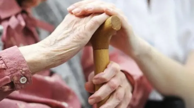 An older person's hand on a cane, being touched by the hand of a younger woman