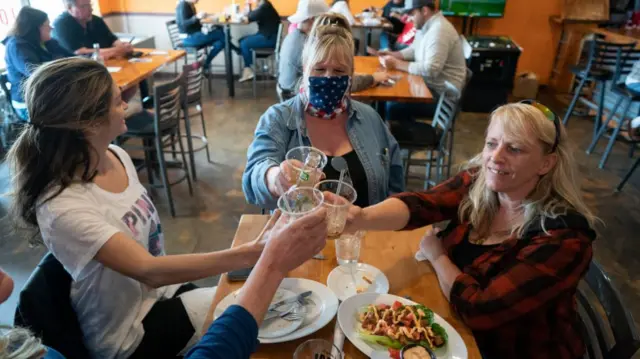 Four people together clinking glasses in a restaurant/bar. One has a face mask.