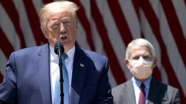 Trump at a podium. Dr Fauci stands behind him wearing a face mask