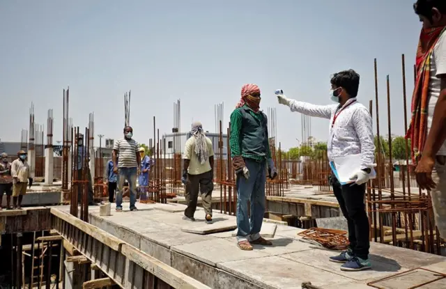 A man holds a temperature gun to another man's head at a building site