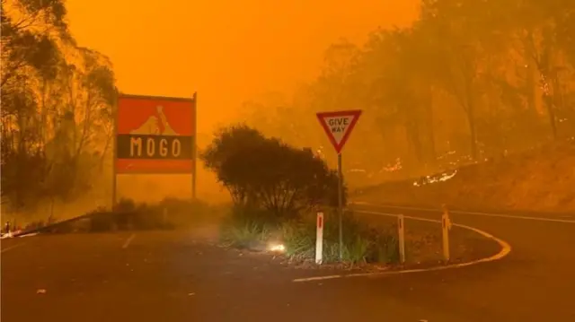 A road sign says "Mogo" as bushfires rage in the background