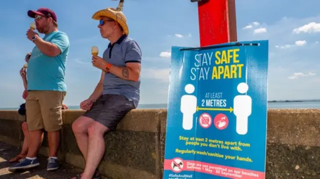 People eating ice creams stand near a sign telling them to social distance at the beach