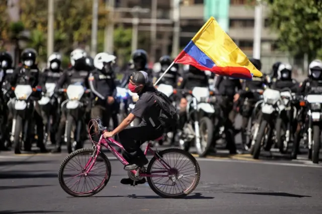 A man riding a bicycle flutters an Ecuadorean flag in front of a line of policemen in downtown Quito