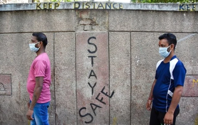 Two Indian men wearing masks and standing in front a wall that says Stay Safe in paint