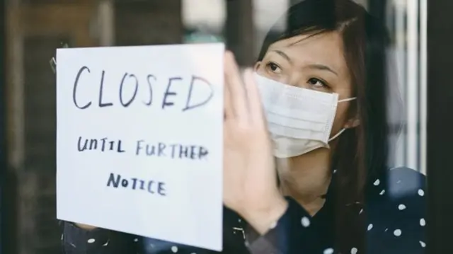 A woman wearing a mask puts up a 'Closed' sign in a window