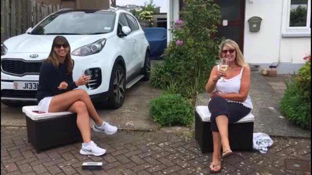 Two women sit at a physical distance from each other drinking wine outside their homes