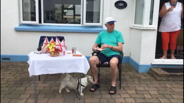 Man eats cake at a table in his front garden, with dog nearby