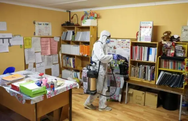 A cleaner wearing a hazmat suit and mask cleans a classroom