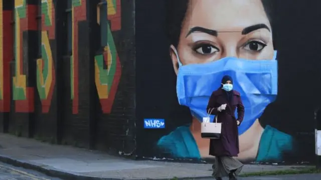 A woman wearing a face mask walks past a mural of an NHS worker