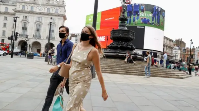 Two people wearing masks and walking across Piccadilly Circus