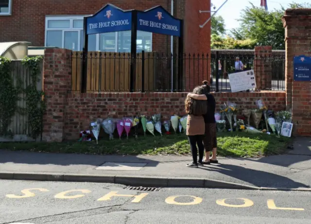 Two people hugging outside The Holt School, next to tributes to James Furlong