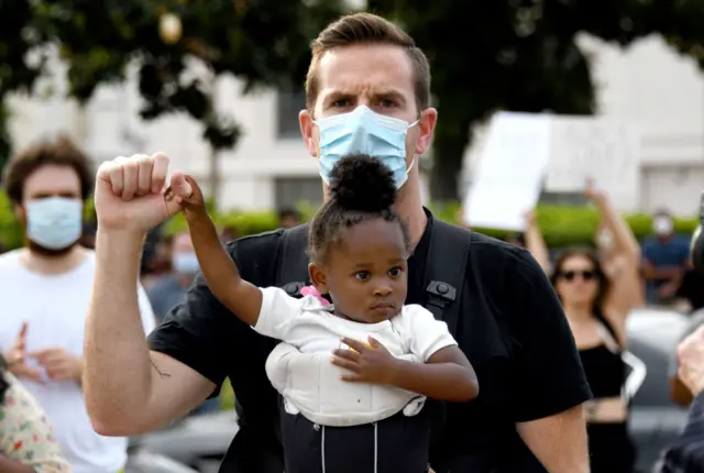 Eric Puestow of Pasadena with his 1 1/2 year-old daughter Simone holds his fist up as she holds onto his hand as thousands of demonstrators caravaned to the Pasadena City Hal