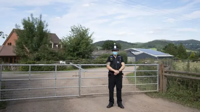 A police officer wearing a surgical face mask stands at the entrance to AS Green and Co farm