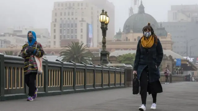 Two women wearing masks walk metres apart from each other across a city bridge in Melbourne