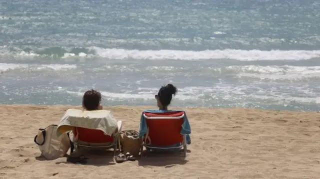 Two people sitting on sunloungers on a beach looking out to sea