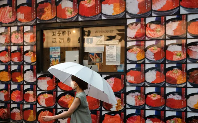 A woman wearing a face mask walks past pictures of sashimi displayed on the side of a restaurant on June 30, 2020 in Tokyo, Japan.