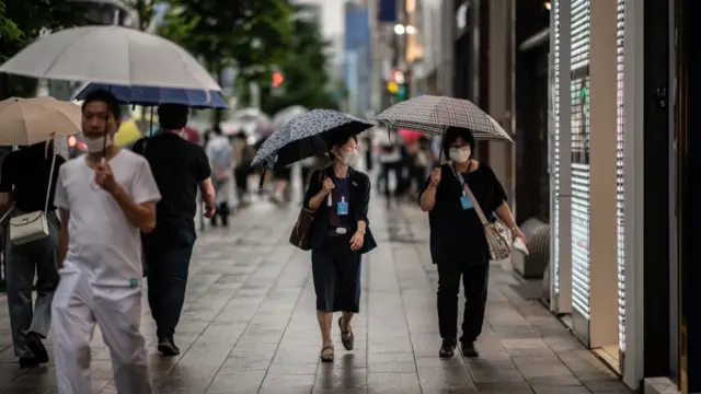 Pedestrians in Tokyo wearing masks and carrying umbrellas, the light is fading.