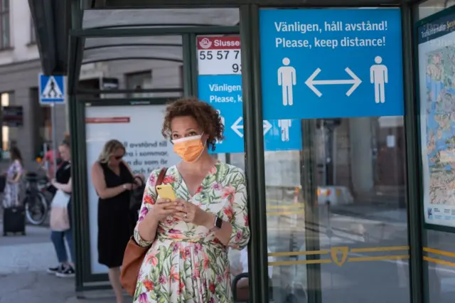 A woman wearing a face mask stands at a bus stop in Stockholm featuring a sign reminding passengers to maintain a minimum social distance between each other to reduce the risk of infection (June 2020)