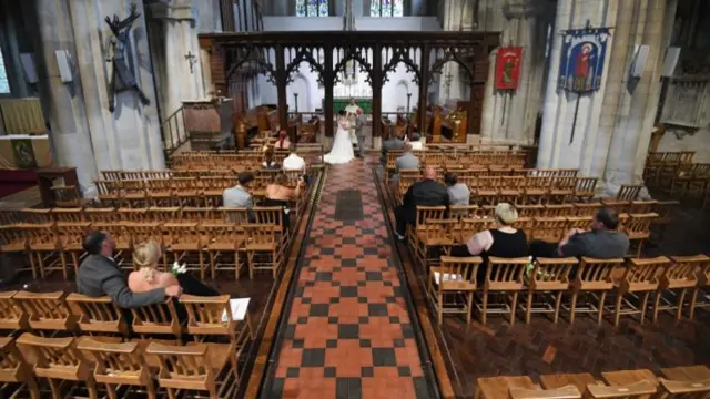 Pairs of socially distanced people sitting in every other row of chairs inside a church watch two people get married