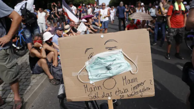 A cardboard sign shows a face wearing a mask and says: "Oh man, how long shall this be going on like this?"