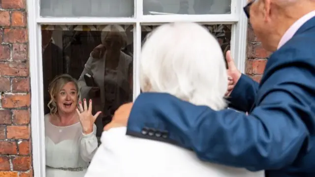 A bride shows off her wedding ring to her parents through a window