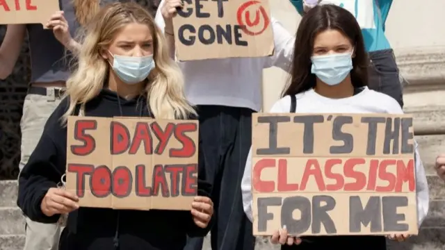 Students in masks hold cardboard placards which read "5 days too late" and "it's the classism for me"