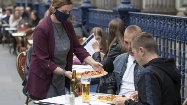 Two men being served a pizza by a waitress wearing a mask