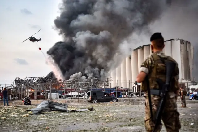 Lebanese army soldiers stand while behind a helicopter puts out a fire at the scene of an explosion at the port of Lebanon's capital Beirut