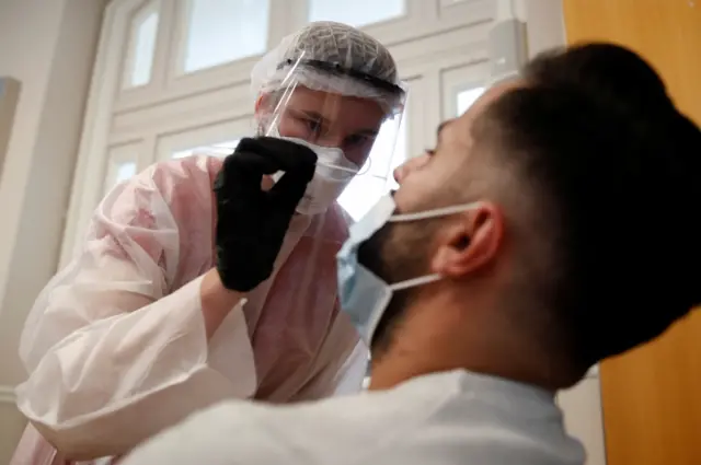 A health worker, wearing a protective suit and a face mask, prepares to administer a nasal swab to a patient