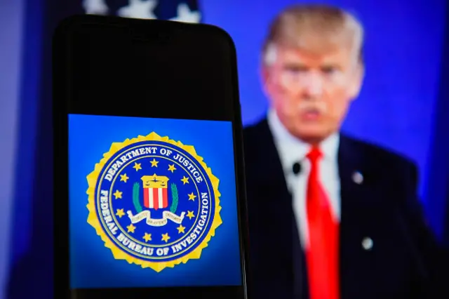 In this photo illustration, the Federal Bureau of Investigation (FBI) logo is seen displayed on an Android mobile phone with the President of the United States of America, DOnald Trump in the background