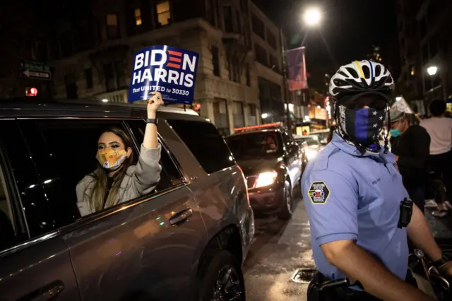 A woman holds a Biden-Harris campaign sign as she passes a police officer during celebrations after President-elect Joe Biden addressed the nation on November 07