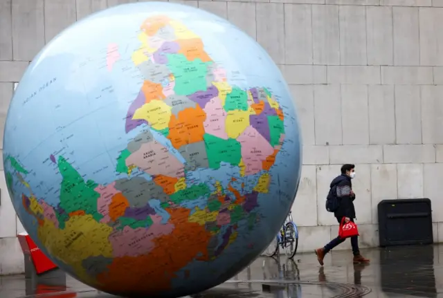 A man wearing a face mask walks past The World Turned Upside Down sculpture by Mark Wallinger