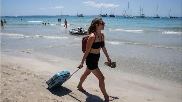 A holidaymaker carries a suitcase across a beach