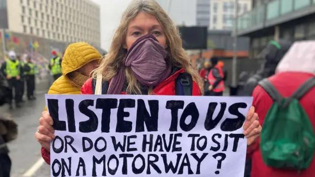 A woman holds up a placard saying 'Listen to us or do we have to sit on a motorway?'