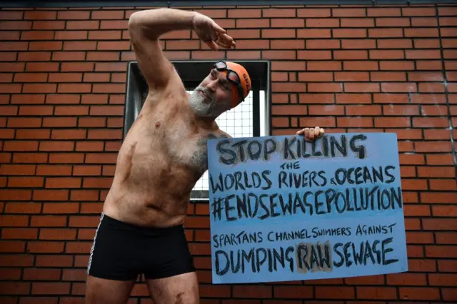 A man who has swam the English Channel, dressed only in swimming trunks, hat and goggles holds up a placard