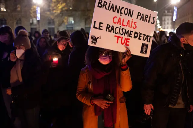 A woman holds a sign that translates as "Briançon, Paris, Calais, the racism of the state kills" at a memorial