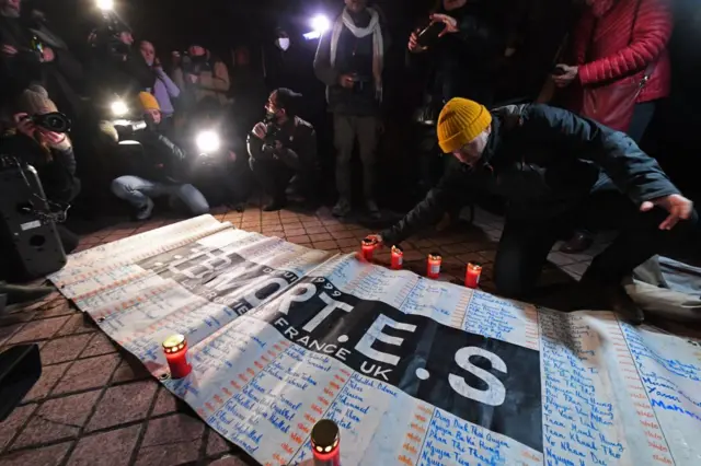 Members of migrant support associations gather next to a placard reading the number of dead and the names of the missing in Calais