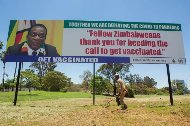 A man cuts grass in front of a billboard with Zimbabwe's President Emmerson Mnangagwa encouraging people to be vaccinated