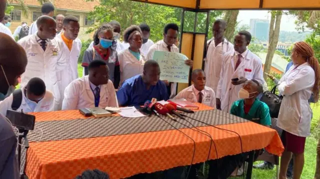 A group of medical staff gathered around a table with a microphones and a protest sign