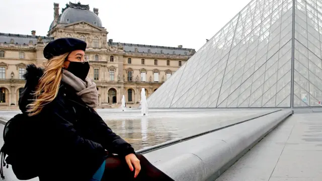 A person wearing a mask sits near the Louvre in Paris