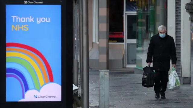 A man in a face mask holding his shopping walks past a 'Thank you NHS' sign