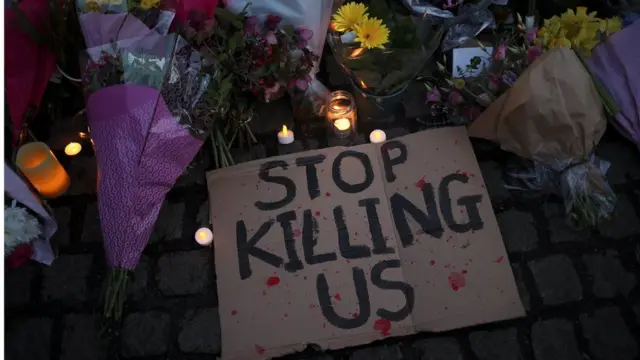 Floral tributes and sign reading: "Stop killing us"