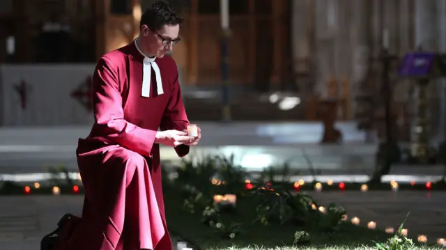 Reverend Canon Andrew Trenier holds a candle next to a green meadow cross adorned with daffodils and spring flowers and surrounded by candles, in the Nave of Winchester Cathedral, ahead of the National Day of Reflection to remember those who have died during the pandemic on 23rd March.