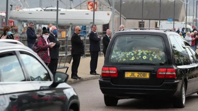 A funeral hearse and onlookers showing their respects in masks