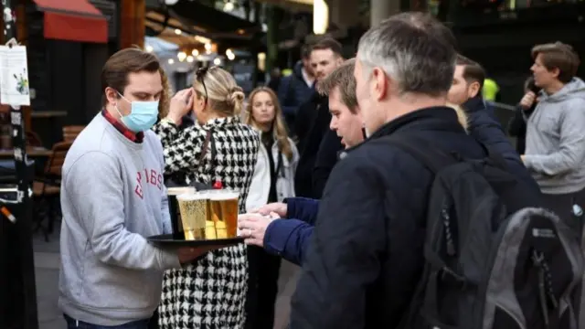 A bartender in a mask serves beer to punters outdoors