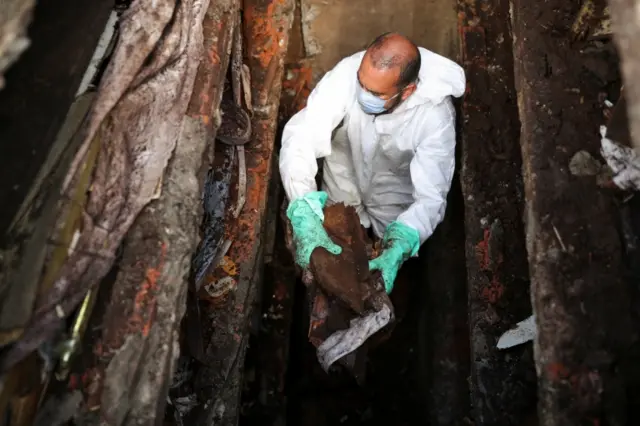 A gravedigger wearing a protective suit handles pieces of an old damaged coffin during exhumations to open space on cement graves